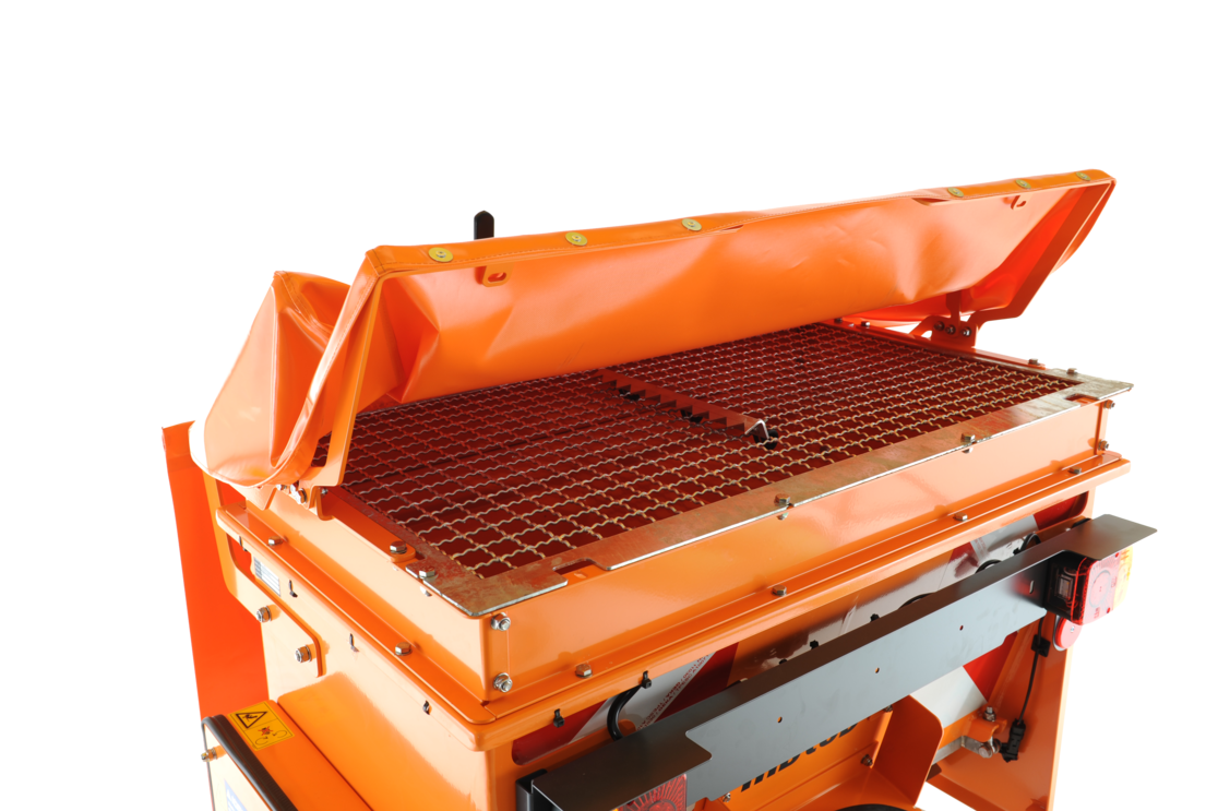 Detailaufnahme eines matev-Kastenstreuers mit geöffneter orangefarbener Abdeckplane. Ein feinmaschiges Schutzgitter bedeckt den Innenbereich, um Fremdkörper fernzuhalten. Im Hintergrund sind Beleuchtungselemente und ein robuster Metallrahmen sichtbar. Die hochwertige Verarbeitung unterstreicht die professionelle Ausrichtung für den Einsatz in der Winter- und Flächenpflege.