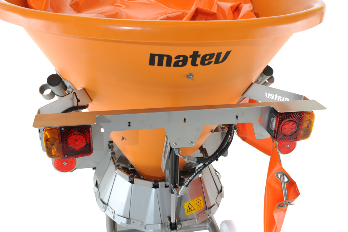 Ein orangefarbener Schleuderstreuer der Marke matev, ausgestattet mit einem stabilen Metallrahmen. Der Streuer verfügt über eine integrierte Beleuchtungseinheit mit Rücklichtern und Reflektoren. Im unteren Bereich sind hydraulische Komponenten und Sicherheitsaufkleber sichtbar. Das Design deutet auf eine professionelle Nutzung im kommunalen oder landwirtschaftlichen Bereich hin.