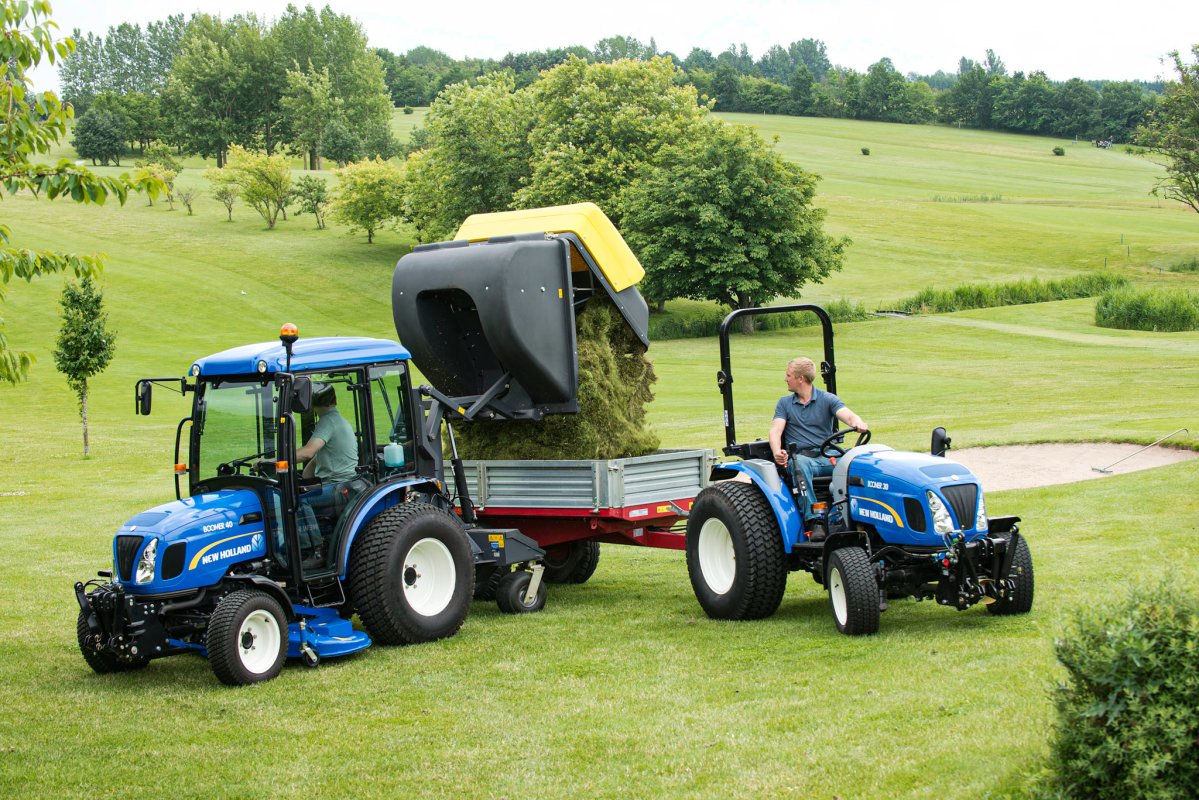 Zwei New Holland Traktoren auf einer gepflegten Grünfläche: Ein Traktor mit matev Grasaufnahmegerät entlädt geschnittenes Gras in einen Anhänger, während der andere Traktor daneben steht. Perfekte Lösung für professionelle Grünflächenpflege in weitläufiger, landschaftlicher Umgebung.