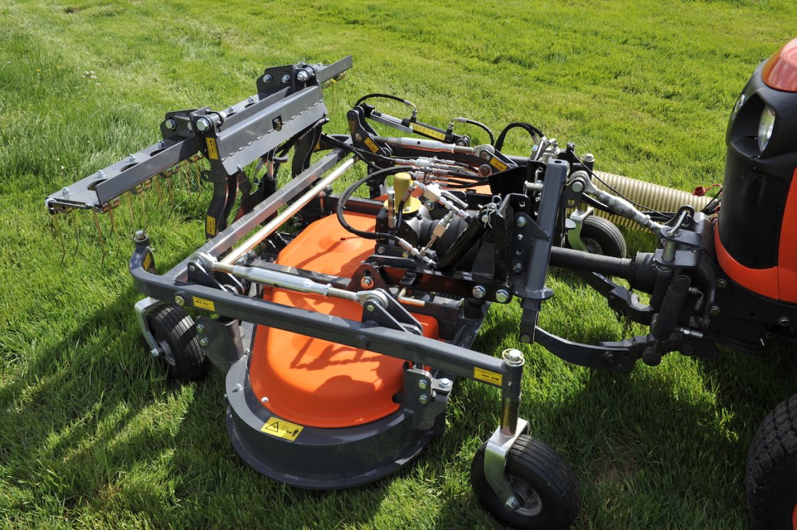 Nahaufnahme eines Matev Frontmähwerks, montiert an einem Kubota STW40 Traktor. Das Mähwerk zeigt komplexe hydraulische Anschlüsse und robuste Mechanik auf einer grünen Wiese.