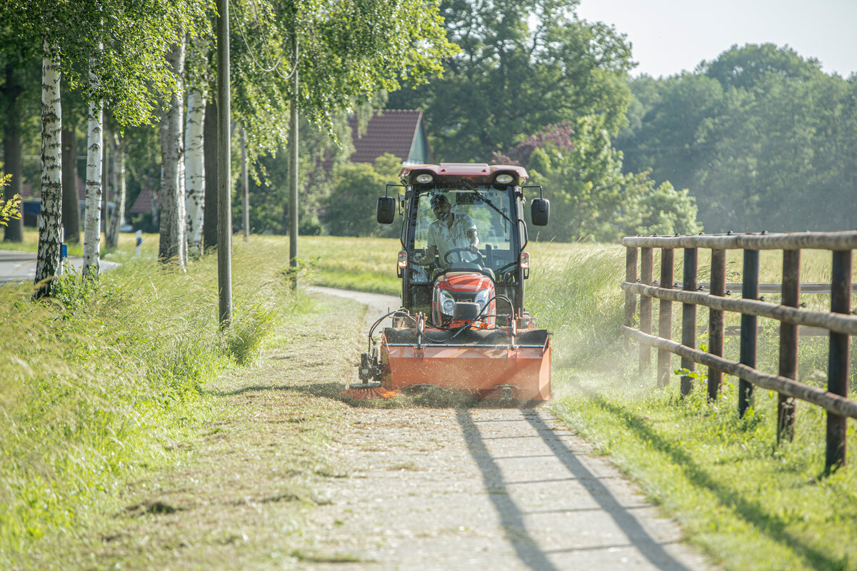 Kioti CX2510 Traktor mit matev Kehrmaschine bei der Reinigung eines schmalen Weges in einer ländlichen Umgebung. Baumreihe und Holzzaun im Hintergrund, ideale Lösung für die professionelle Wegepflege.