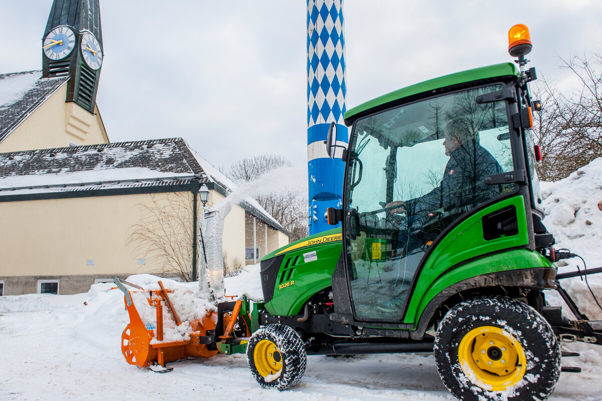 John Deere Traktor 1026R mit angebauter orangefarbener matev-Schneefräse im Einsatz. Die Schneefräse wirft Schnee seitlich aus. Im Hintergrund sind eine Kirche mit Uhrturm, ein traditioneller Maibaum und schneebedeckte Flächen sichtbar. Der Fahrer sitzt in der beheizten Kabine, und die Umgebung zeigt eine winterliche Dorfkulisse.