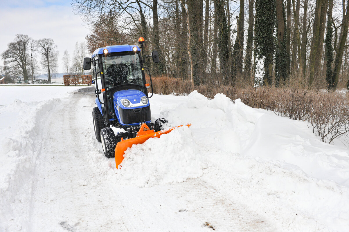Ein blauer New Holland Traktor mit einem orangefarbenen Schneeräumschild von matev räumt eine verschneite Straße frei. Im Hintergrund sind Bäume, Sträucher und schneebedeckte Landschaften zu sehen.