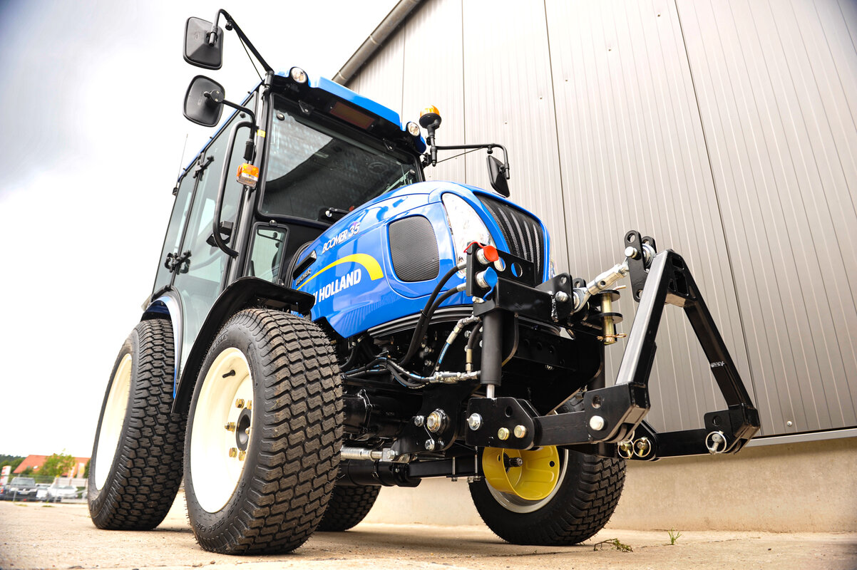 Ein New Holland Boomer 35 Traktor aus der Froschperspektive fotografiert. Der Traktor ist mit einem matev Frontkraftheber ausgestattet, und im Hintergrund ist eine moderne graue Metallhalle zu sehen.