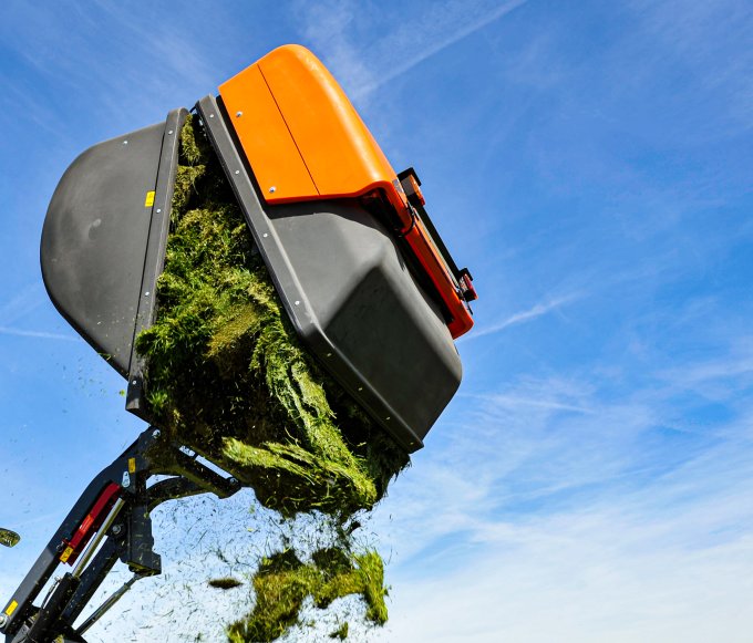Kubota STW40 Traktor mit angebautem Grasaufnahmegerät von matev, das geschnittenes Gras auf ein Feld entlädt. Perfektes Zusammenspiel von Effizienz und Leistung bei der Rasenpflege vor einem klaren blauen Himmel und ländlicher Kulisse.