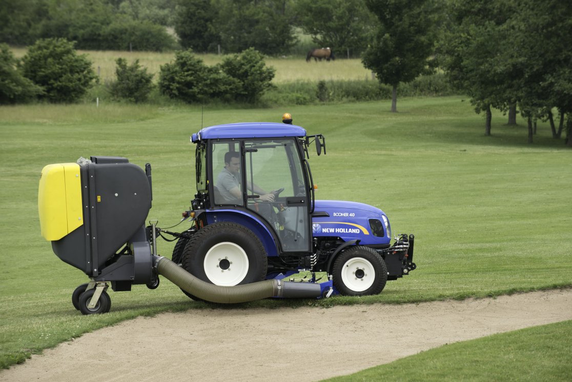 Ein blauer New Holland Boomer 40 Traktor mit einer angehängten matev-Aufnahmeeinheit arbeitet auf einem Golfplatz. Der Traktor sammelt Sand und andere Materialien aus einem Bunker mithilfe eines flexiblen Saugschlauchs, der mit dem gelb-grauen Aufnahmegerät verbunden ist. Im Hintergrund erstreckt sich eine weitläufige Rasenfläche mit Bäumen und einem Pferd auf einer Wiese. Die Szene zeigt eine professionelle Anwendung in der Platzpflege.