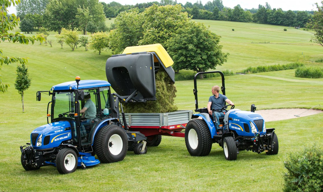 Zwei New Holland Traktoren auf einer gepflegten Grünfläche: Ein Traktor mit matev Grasaufnahmegerät entlädt geschnittenes Gras in einen Anhänger, während der andere Traktor daneben steht. Perfekte Lösung für professionelle Grünflächenpflege in weitläufiger, landschaftlicher Umgebung.