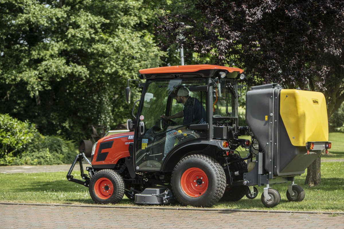 Ein Kioti CX2510 Traktor mit Matev Multifunktionsmähwerk und gelbem Materialaufnahmegerät im Einsatz auf einer gepflegten Rasenfläche. Der Fahrer arbeitet konzentriert, umgeben von dichter grüner Vegetation und einem Baum im Hintergrund. | © Daedong Kioti Europe B.V.