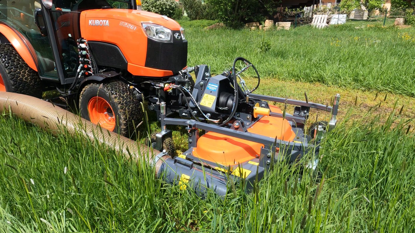 Nahaufnahme eines Kubota B2261 Traktors mit einem Matev Frontmähwerk und Grasaufnahmegerät, das hohes Gras auf einer Wiese mäht. Der Schlauch für die Grasaufnahme ist gut sichtbar, umgeben von saftigem Grün.