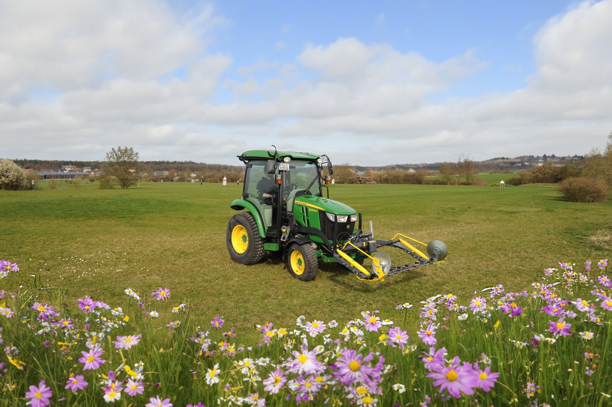 Ein grüner John Deere 3045R Traktor mit einem matev MOW-PT Portalmähwerk steht auf einer grünen Wiese. Im Vordergrund blühen lila und gelbe Wildblumen, während im Hintergrund eine ländliche Landschaft mit Bäumen, Gebäuden und Hügeln unter blauem Himmel sichtbar ist.