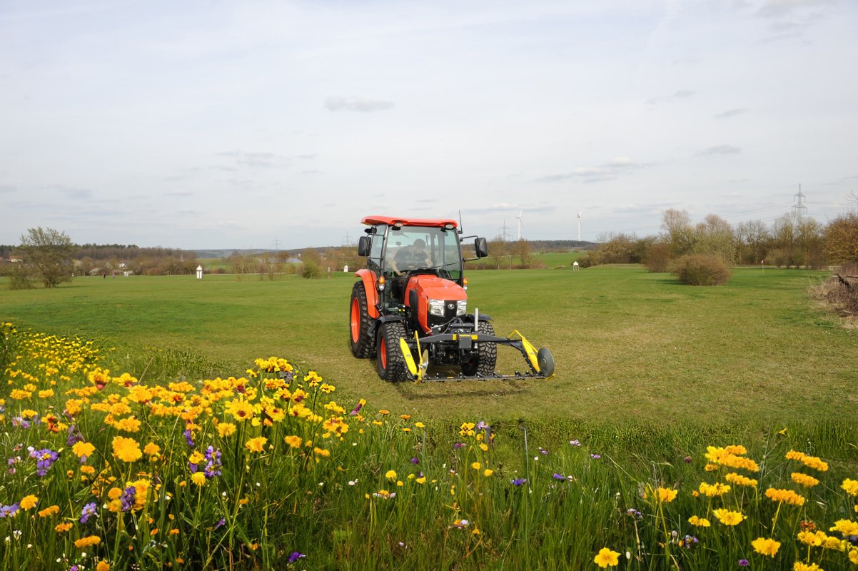 Ein roter Kubota L2-622 Traktor mit einem matev MOW-PT Portalmähwerk steht auf einer grünen Wiese. Im Vordergrund blühen gelbe und lila Wildblumen, während im Hintergrund eine ländliche Landschaft mit Windrädern, Bäumen und einem Pferd sichtbar ist.