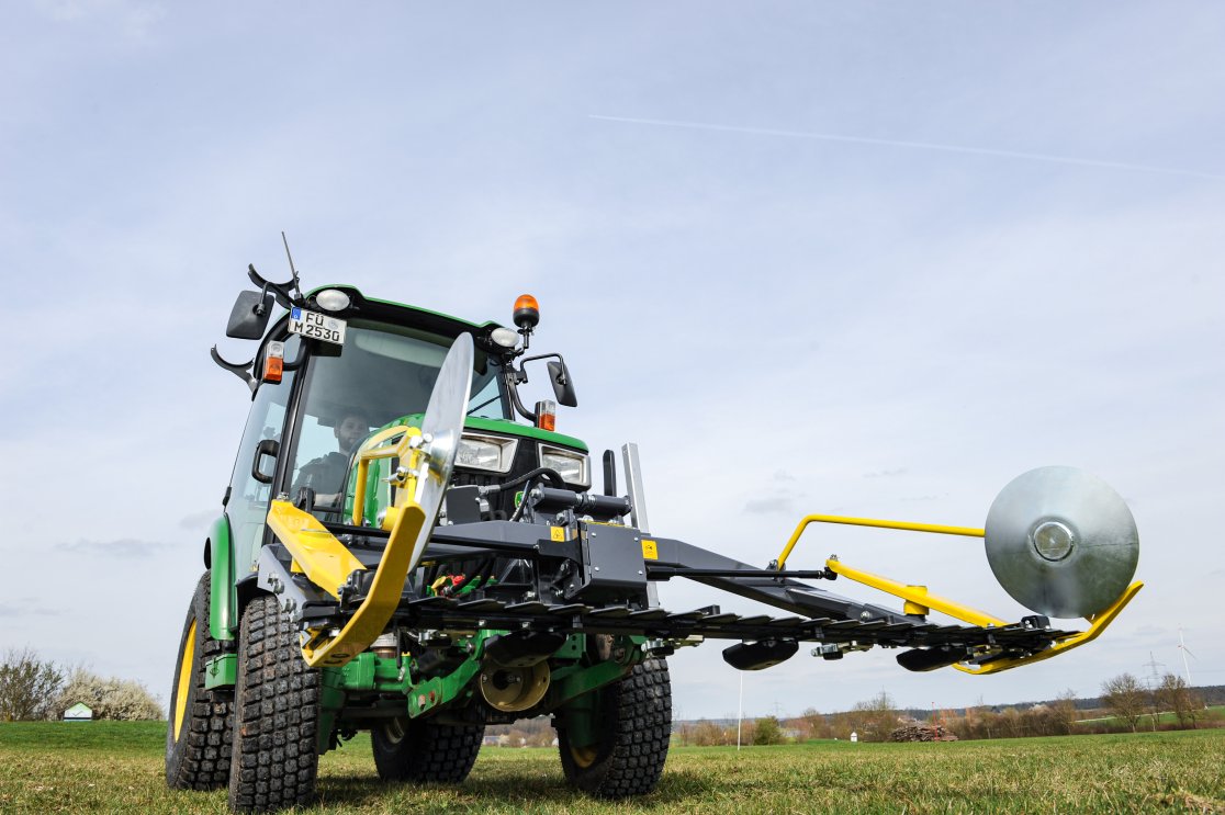 Ein grüner John Deere Traktor der 3R-Serie mit einem matev MOW-PT Portalmähwerk, fotografiert aus einer niedrigen Perspektive. Das gelb-graue Mähwerk ist im Vordergrund hervorgehoben, während im Hintergrund eine ländliche Landschaft mit Bäumen und Windrädern zu sehen ist.