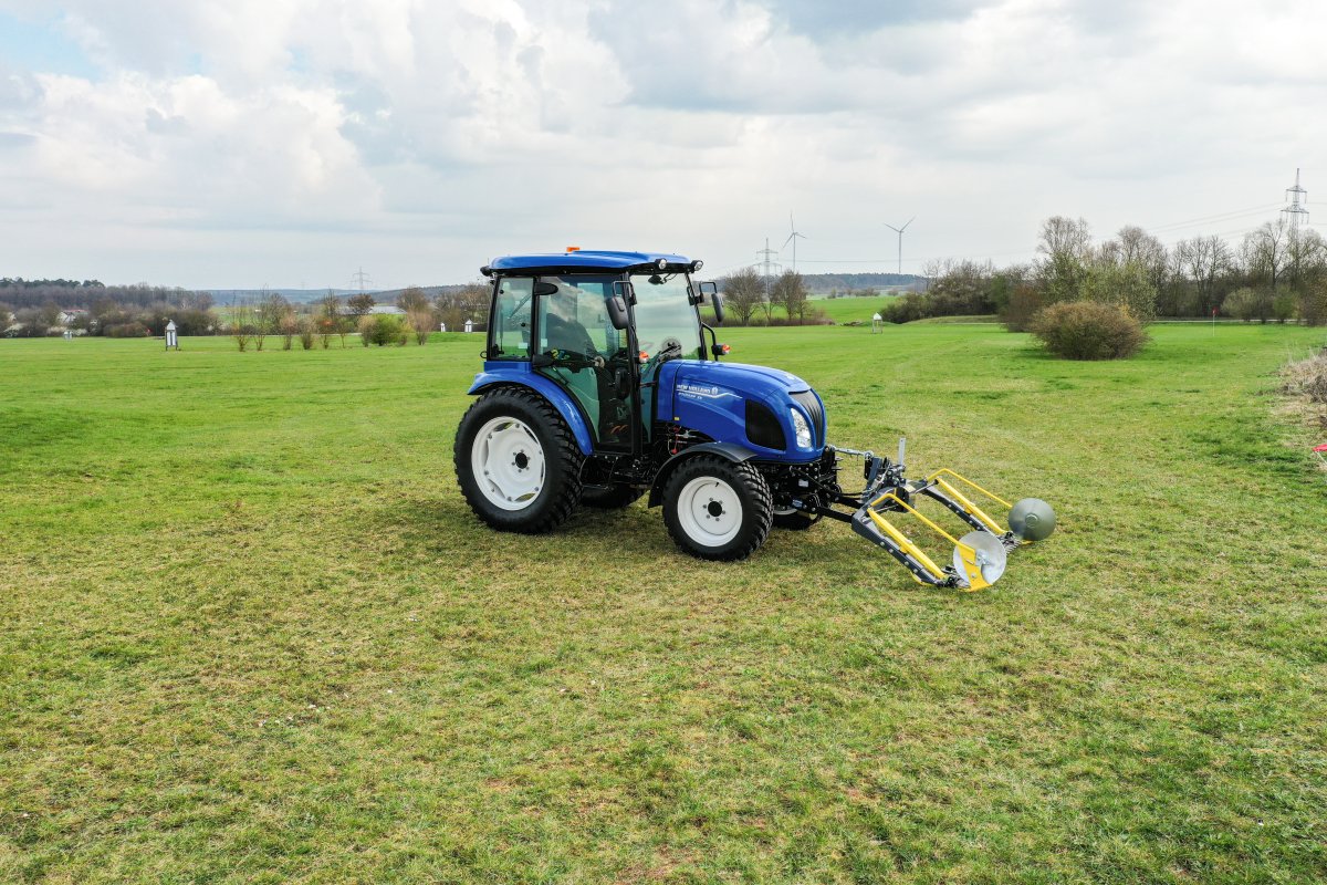 Ein blauer New Holland Boomer 55 Traktor mit Kabine und einem matev MOW-PT Portalmähwerk steht auf einer weitläufigen grünen Wiese. Im Hintergrund sind Bäume, Windräder und Strommasten in einer ländlichen Umgebung zu sehen.