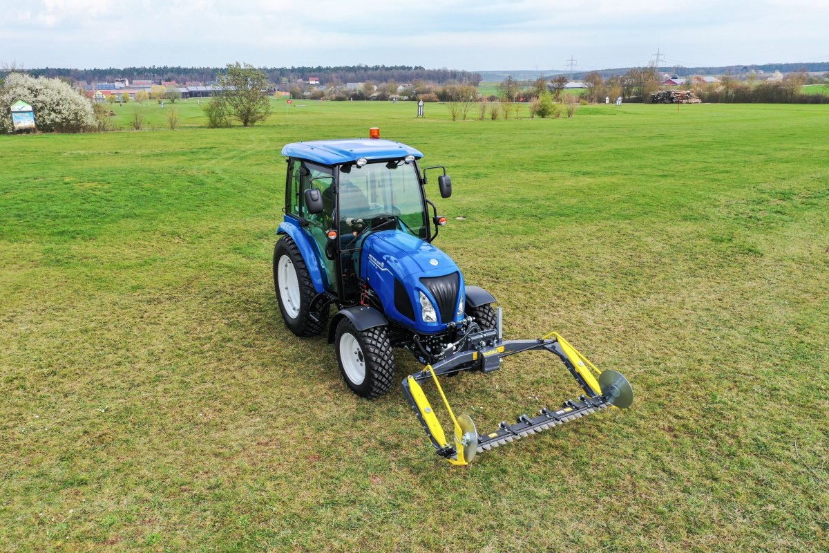 Ein blauer New Holland Boomer 55 Traktor mit einem angebauten matev MOW-PT Portalmähwerk steht auf einer großen grünen Wiese. Im Hintergrund erstreckt sich eine ländliche Umgebung mit Gebäuden, Bäumen und Strommasten.