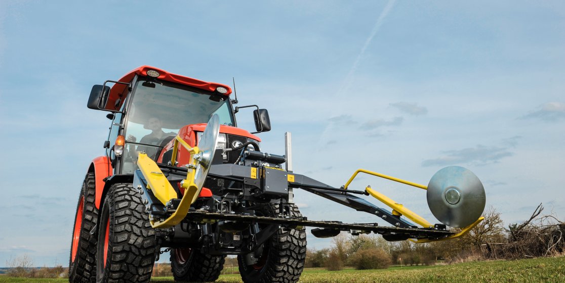 Ein roter Kompakttraktor, ausgestattet mit einem matev MOW-PT Portalmähwerk, steht auf einer grünen Wiese unter klarem Himmel. Die gelb-schwarze Konstruktion des Mähwerks ist prominent im Vordergrund sichtbar und betont die präzise Technik.
