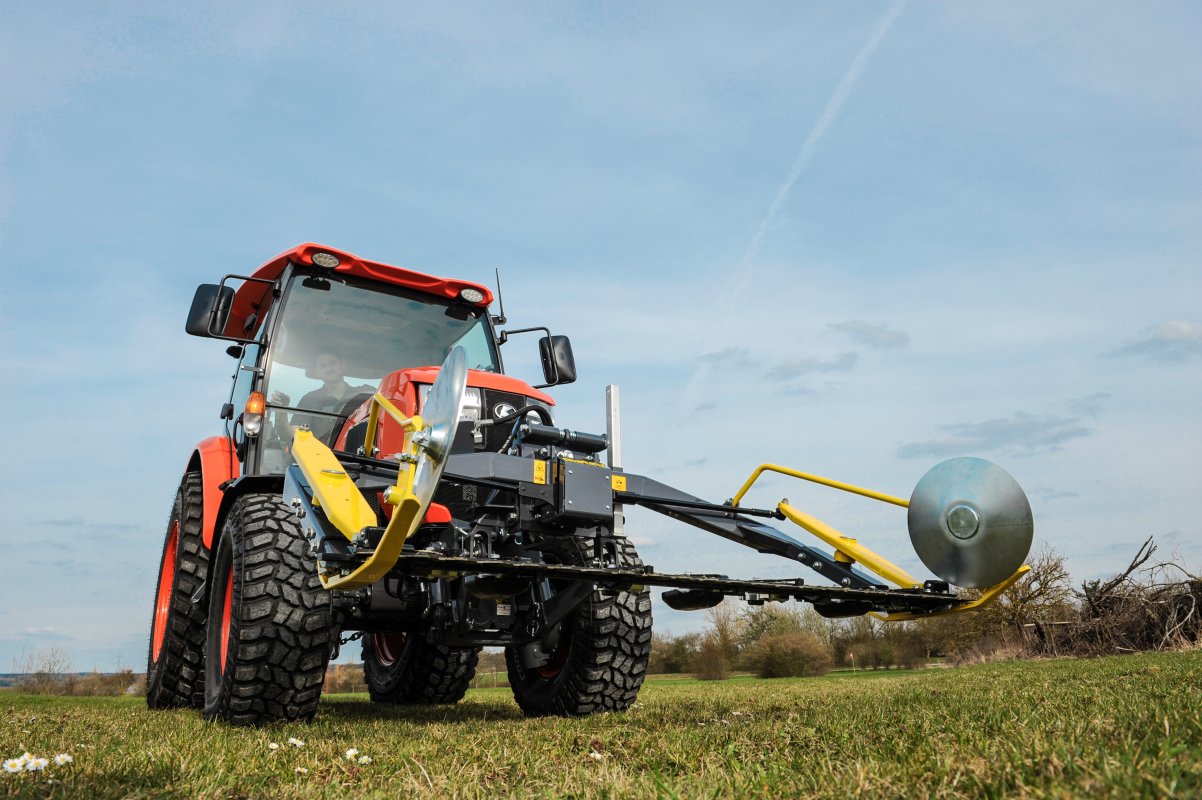 Ein roter Kompakttraktor, ausgestattet mit einem matev MOW-PT Portalmähwerk, steht auf einer grünen Wiese unter klarem Himmel. Die gelb-schwarze Konstruktion des Mähwerks ist prominent im Vordergrund sichtbar und betont die präzise Technik.