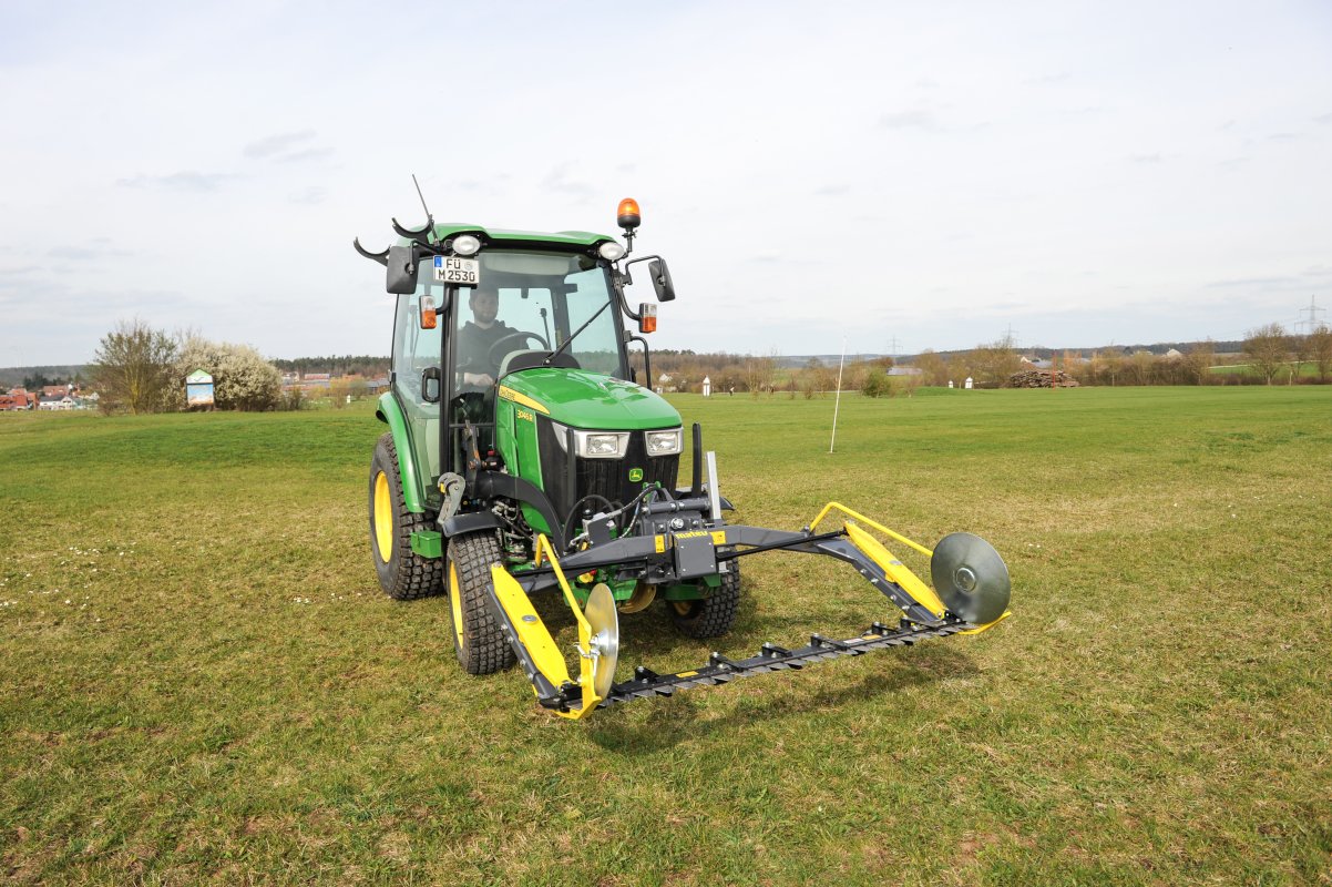 Ein grüner John Deere Traktor der 3R-Serie mit einem matev MOW-PT Portalmähwerk steht auf einer Wiese. Im Hintergrund sind Bäume, ein Dorf und Strommasten zu sehen, die eine ländliche Umgebung darstellen.
