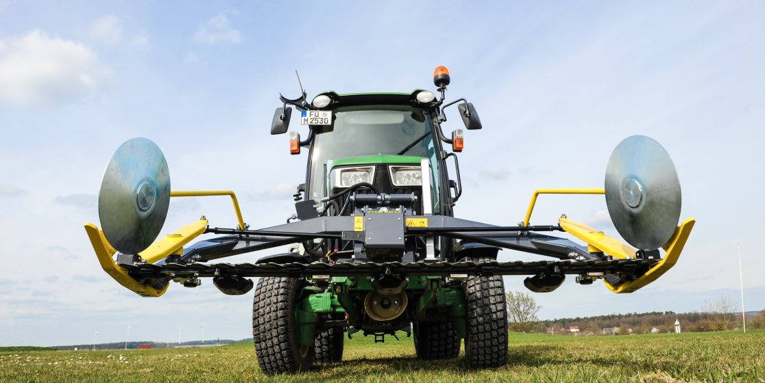 Ein John Deere Traktor der 3R-Serie mit einem Matev Portalmähwerk im Einsatz auf einer Wiese. Das Portalmähwerk ist frontal abgebildet, mit seinen großen gelben Schutzscheiben und klar sichtbarer Technik, unter einem leicht bewölkten Himmel.