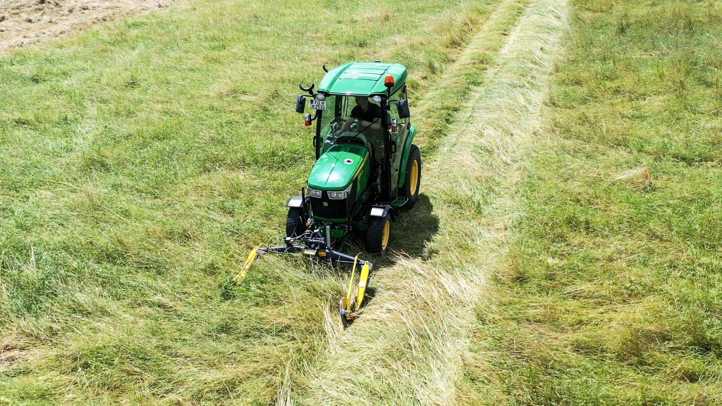 Ein John Deere Traktor der 3R-Serie, ausgestattet mit einem matev MOW-PT Portalmähwerk, mäht eine Wiese und hinterlässt saubere Mähspuren. Die grüne Landschaft und die Struktur des Grases zeigen eine effiziente Mäharbeit.