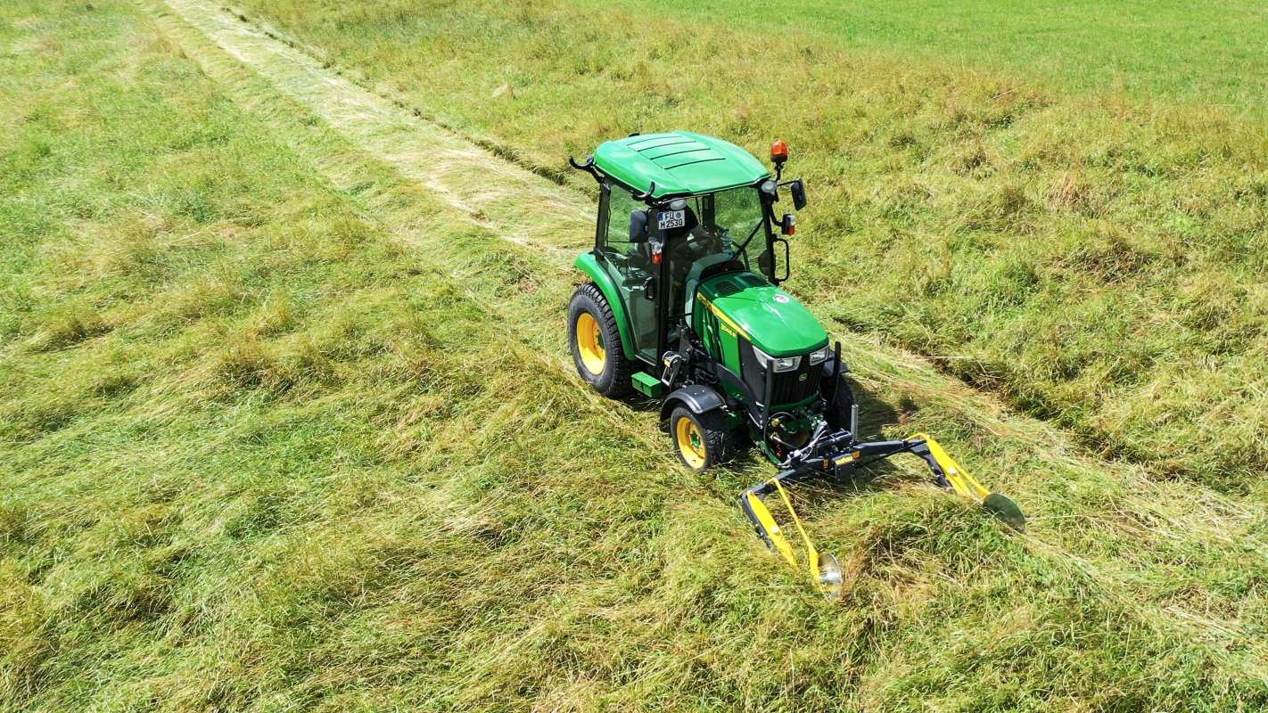 Ein John Deere Traktor der 3R-Serie mit einem matev MOW-PT Portalmähwerk mäht Gras auf einer weitläufigen, grünen Wiese. Die frisch gemähten Spuren heben sich deutlich von der umliegenden Vegetation ab, und die ländliche Landschaft vermittelt einen idyllischen Eindruck.