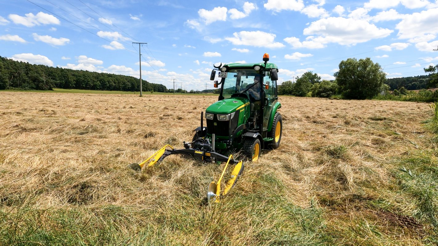 Ein grüner John Deere Traktor der 3R-Serie mit einem matev MOW-PT Portalmähwerk mäht eine große Wiese mit hohem Gras. Im Hintergrund sind Stromleitungen, Bäume und ein klarer Himmel mit vereinzelten Wolken zu sehen.