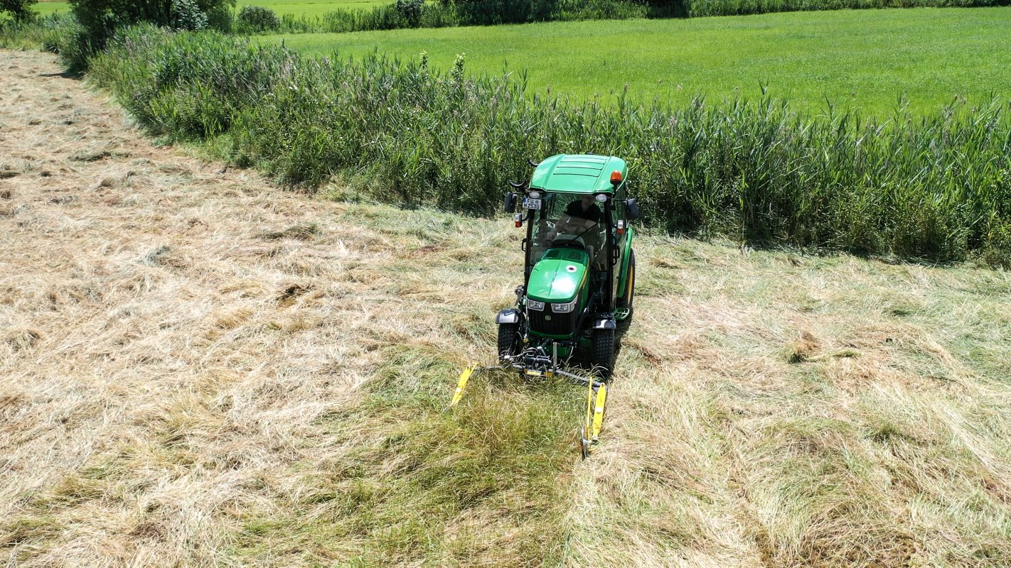 Ein John Deere Traktor der 3R-Serie ist mit einem matev MOW-PT Portalmähwerk ausgestattet und mäht hohes Gras auf einer Wiese. Im Hintergrund sind grüne Felder und ein dichter Schilfgürtel zu sehen, die die natürliche Umgebung unterstreichen.