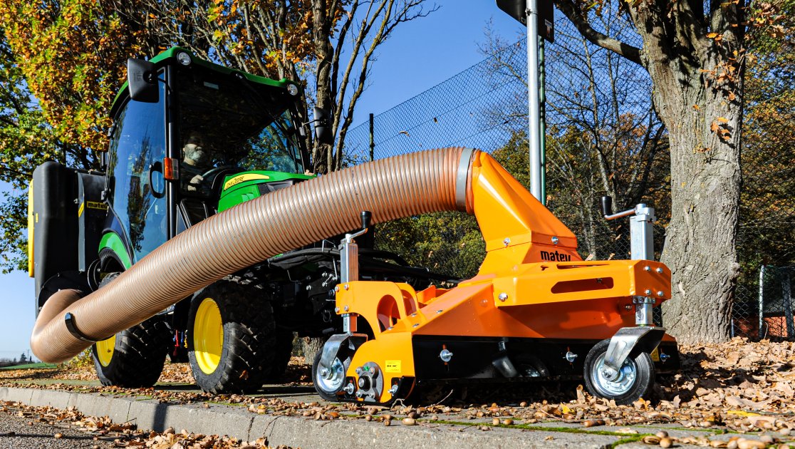 Ein kompaktes Multifunktions-Sauggerät, verbunden mit einem grünen John Deere Kompakttraktor. Das orangefarbene Gerät verfügt über einen flexiblen Saugschlauch und robuste Räder, ideal für die effiziente Aufnahme von Laub und Schmutz in kommunalen oder gartenbaulichen Bereichen.