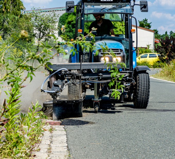 Traktor mit angebauter Wildkrautbürste von matev bei der Arbeit am Straßenrand. Die Bürste entfernt Unkraut entlang einer Bordsteinkante. Im Vordergrund sind hochgewachsene Pflanzen und Wildblumen sichtbar. Der Traktor fährt auf einer asphaltierten Straße, während im Hintergrund Bäume, ein Zaun und ein gelbes Auto zu erkennen sind. Der Himmel ist sonnig und klar.
