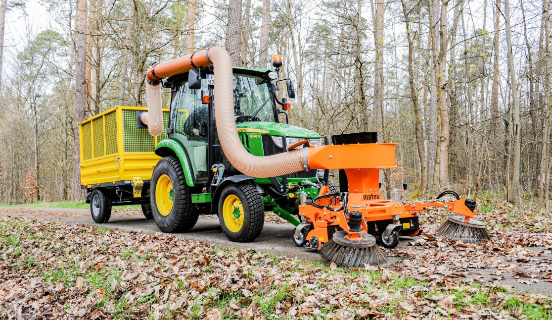Ein Anbaugerät Saugkehrmaschine im Einsatz an einem John Deere 2026r Traktor im Wald zur Flächenpflege und im kommunalen Einsatz.