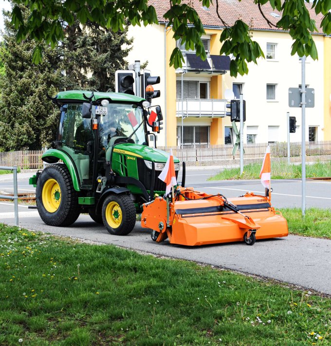 John Deere 3R Traktor mit matev Kehrmaschine in Orange bei der Reinigung eines Gehwegs neben einer Bahnschranke und Wohnhäusern. Perfekte Lösung für die Pflege von Gehwegen und urbanen Flächen.