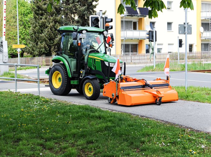 John Deere 3R Traktor mit matev Kehrmaschine in Orange bei der Reinigung eines Gehwegs neben einer Bahnschranke und Wohnhäusern. Perfekte Lösung für die Pflege von Gehwegen und urbanen Flächen.