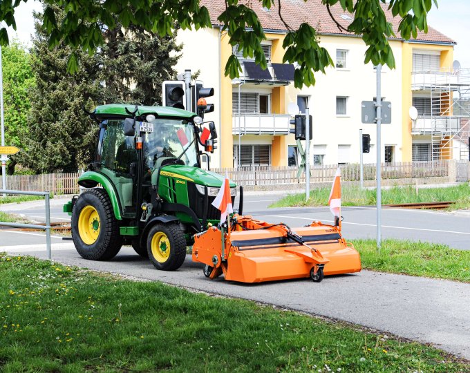 John Deere 3R Traktor mit matev Kehrmaschine in Orange bei der Reinigung eines Gehwegs neben einer Bahnschranke und Wohnhäusern. Perfekte Lösung für die Pflege von Gehwegen und urbanen Flächen.