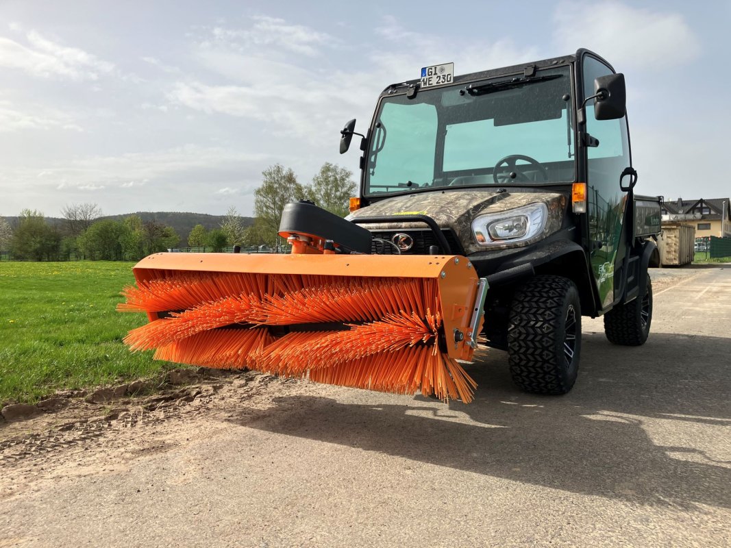 Ein Kubota RTV in Camouflage-Optik mit einer angebauten matev Kehrmaschine SWE 16/45. Die orangefarbenen Bürsten der Kehrmaschine heben sich kontrastreich vom grünen Gras im Hintergrund ab. Das Fahrzeug steht auf einem befestigten Weg mit leicht bewölktem Himmel und umgebender Natur.