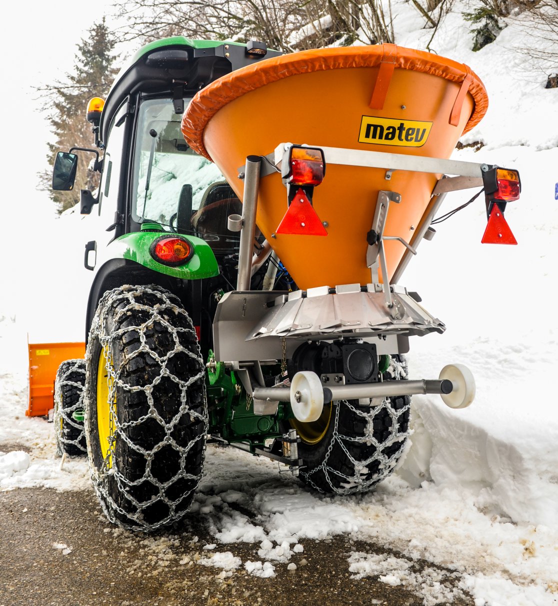 Rückansicht eines John Deere Traktors mit angebautem orangefarbenem matev-Schleuderstreuer DS auf einem schneebedeckten Weg. Die Traktorreifen sind mit Schneeketten ausgestattet, und im Hintergrund befinden sich verschneite Hänge und Bäume. Das orangefarbene Schneeräumschild ist an der Vorderseite des Traktors sichtbar. Die Szene zeigt eine winterliche Umgebung.
