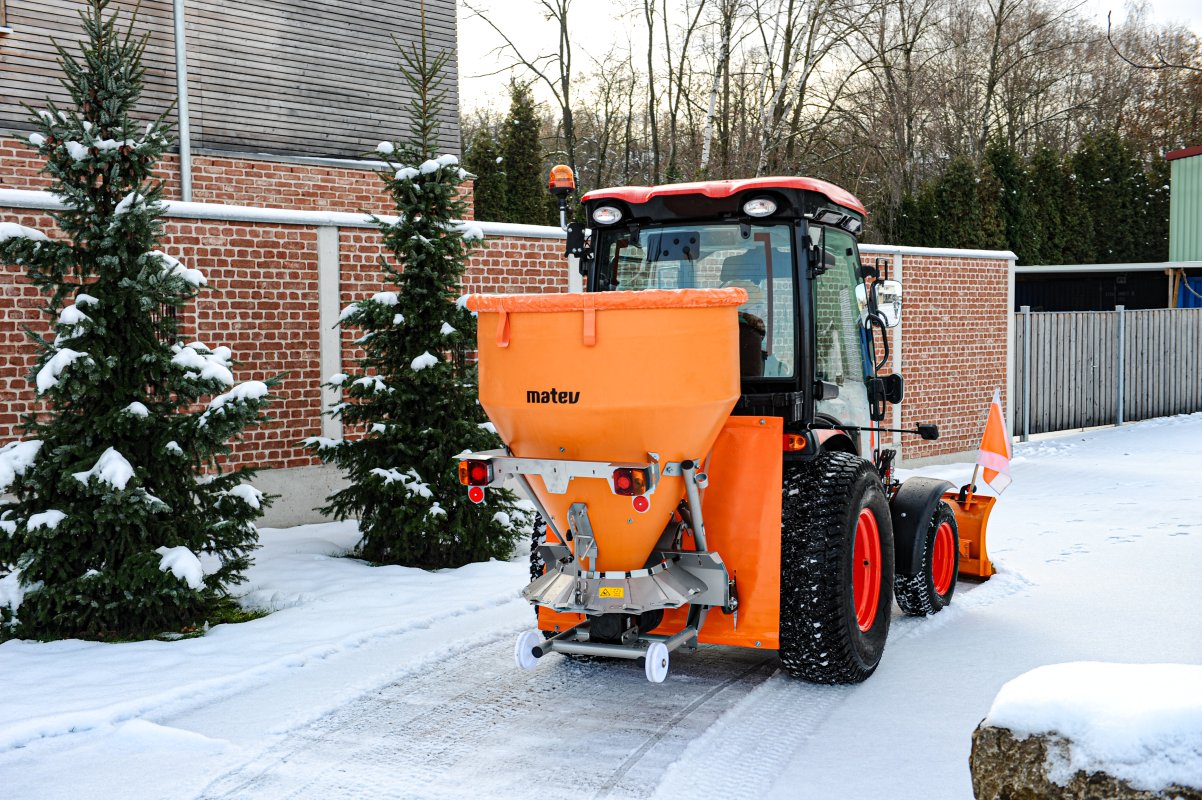 Rückansicht eines Traktors mit angebautem orangefarbenem matev-Tellerstreuer IX-600 und Schneeräumschild. Der Traktor steht auf einem schneebedeckten Weg. Im Hintergrund befinden sich verschneite Tannen, eine Ziegelmauer und ein modernes Gebäude. Der Himmel ist bewölkt, und der Bereich wirkt winterlich gepflegt.