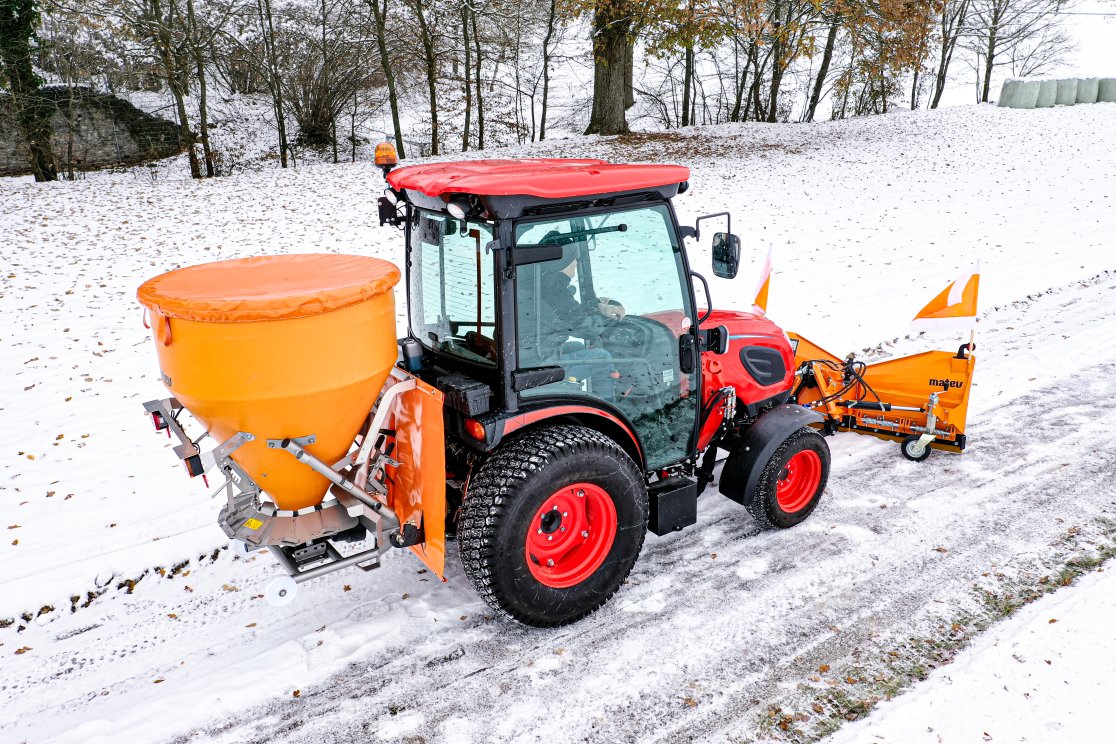 Seitenansicht eines roten Traktors mit angebautem orangefarbenem matev-Tellerstreuer IX-600 und Schneepflug KI-CK4030. Der Traktor fährt auf einer schneebedeckten Straße, die von einer winterlichen Landschaft mit Bäumen und Laub im Schnee umgeben ist. Der Fahrer ist in der Kabine sichtbar, und der Streuer ist mit einer Abdeckplane ausgestattet.