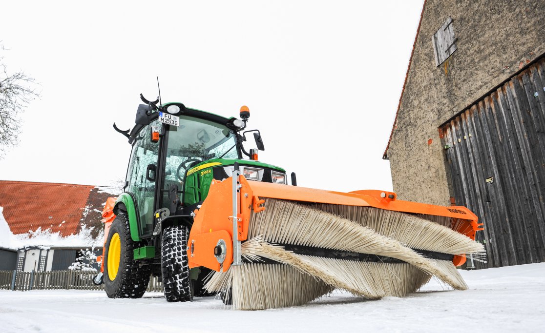 John Deere 3046R Traktor mit matev Kehrmaschine SWE-57 und Streuer SPR-BX-100 im Einsatz auf verschneitem Hof, ideal für Winterdienst und Schneeräumung.
