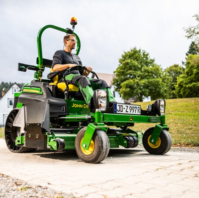 Ein Mann fährt auf einem John Deere Z997R Aufsitzmäher mit StVZO-Kit von matev. Das Fahrzeug ist auf einem gepflasterten Weg in einer grünen, ländlichen Umgebung zu sehen. Im Hintergrund stehen Häuser, Bäume und Hügel.