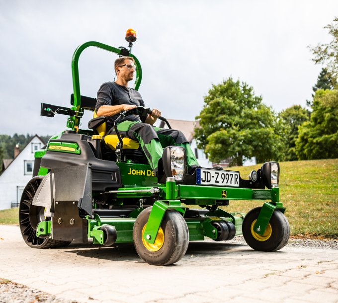 Ein Mann fährt auf einem John Deere Z997R Aufsitzmäher mit StVZO-Kit von matev. Das Fahrzeug ist auf einem gepflasterten Weg in einer grünen, ländlichen Umgebung zu sehen. Im Hintergrund stehen Häuser, Bäume und Hügel.
