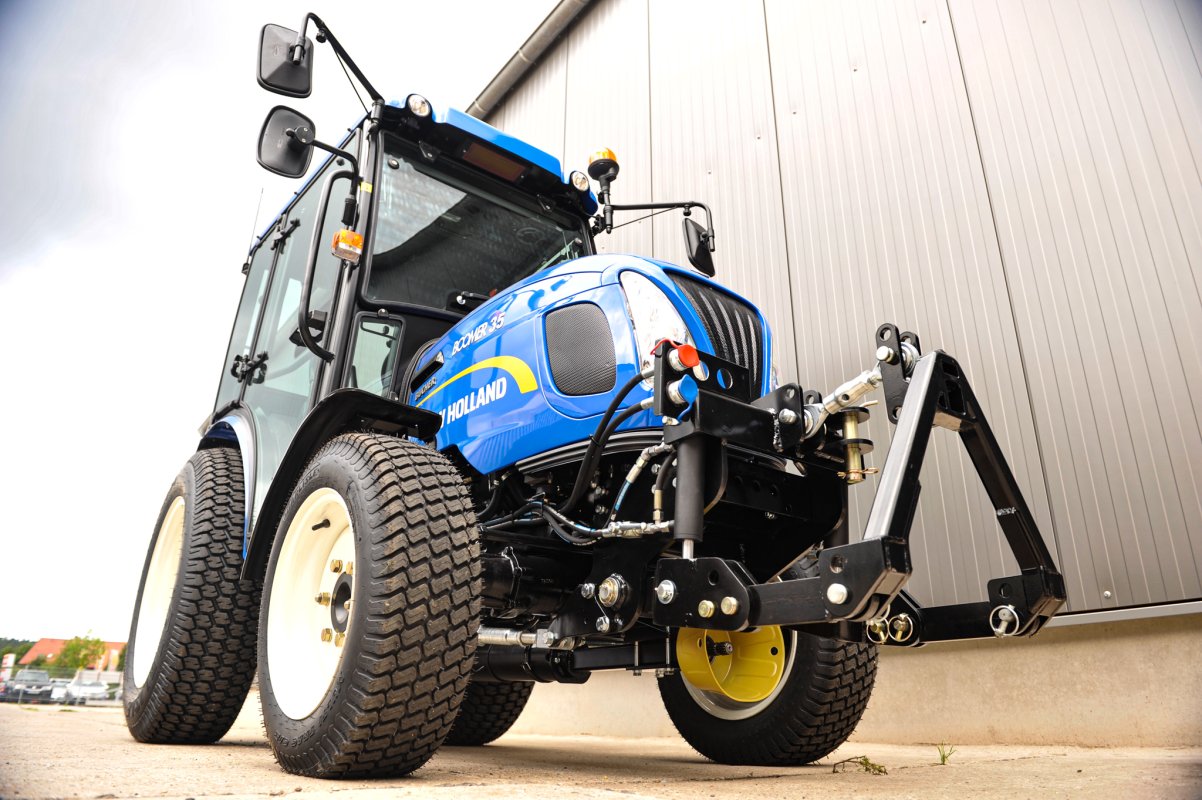 Ein New Holland Boomer 35 Traktor aus der Froschperspektive fotografiert. Der Traktor ist mit einem matev Frontkraftheber ausgestattet, und im Hintergrund ist eine moderne graue Metallhalle zu sehen.