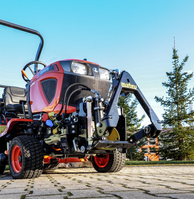 Roter Kubota Kompakttraktor Modell G231, ausgestattet mit einem matev Frontkraftheber, fotografiert aus einer niedrigen Perspektive auf gepflastertem Untergrund, umgeben von Bäumen und blauem Himmel.
