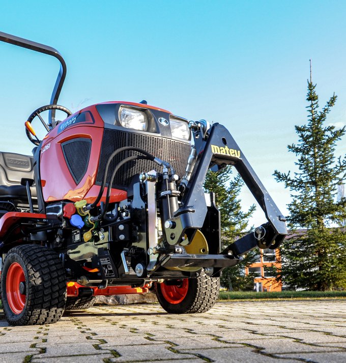 Roter Kubota Kompakttraktor Modell G231, ausgestattet mit einem matev Frontkraftheber, fotografiert aus einer niedrigen Perspektive auf gepflastertem Untergrund, umgeben von Bäumen und blauem Himmel.