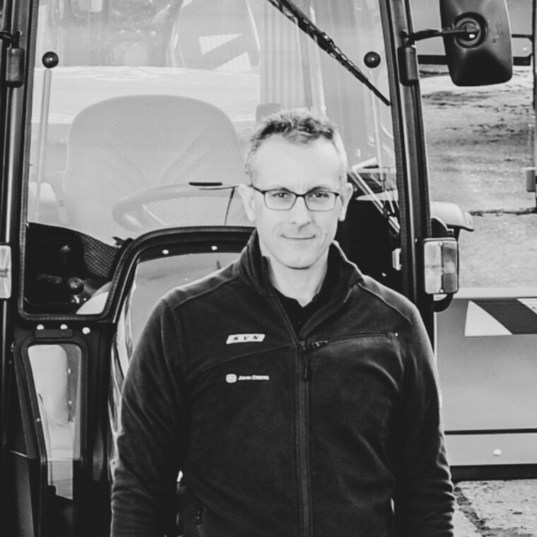 Schwarz-weiß Porträt von Frank Weidenberg der KVN in Nürnberg mit Brille und Jacke, stehend vor der Fahrerkabine eines Traktors, der professionell und freundlich in die Kamera schaut.
