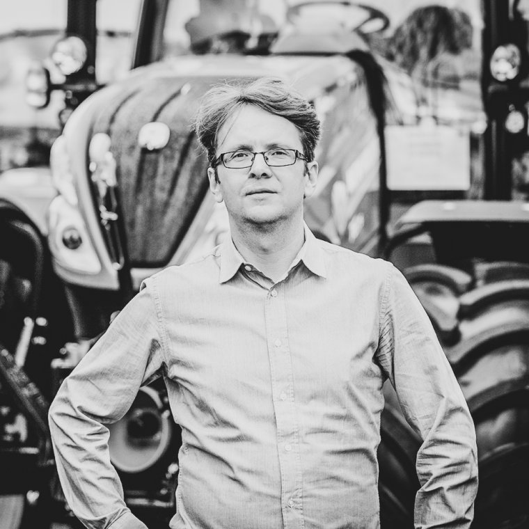 Schwarz-weiß Porträt von Leo Cermak von Agrotec mit Brille und Hemd, stehend vor einem Traktor, der den Hintergrund ausfüllt. Der Mann blickt selbstbewusst in die Kamera.