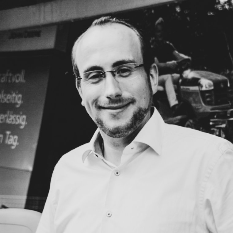 Schwarz-weiß Porträt von Christian Buchard von der LVN Steinbrink mit Brille und Bart in einem Hemd, freundlich lächelnd, vor einem Hintergrund mit John Deere-Marketingmaterial und einem Traktor."