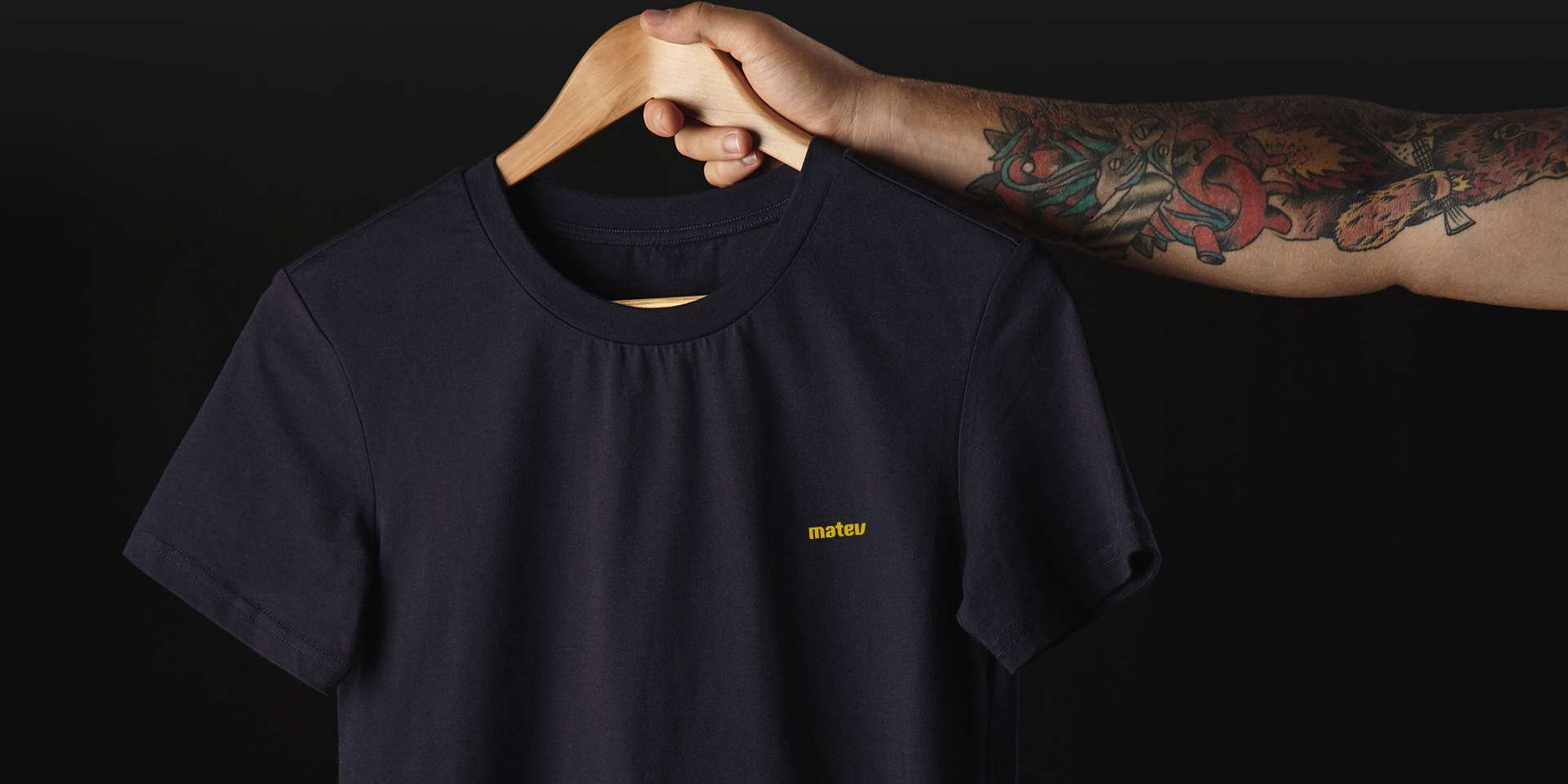 Schwarzes T-Shirt mit gelbem matev-Logo auf einem Holzkleiderbügel, gehalten von einem tätowierten Arm vor einem schwarzen Hintergrund.