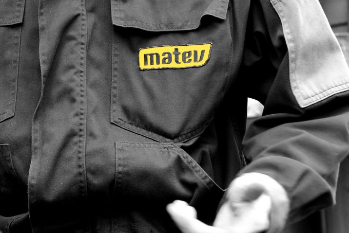 Schwarz-weißes Bild eines Mitarbeiters in Arbeitskleidung mit dem gelben matev-Logo auf der Brusttasche der Jacke, Fokus auf Details der robusten Stoffstruktur.